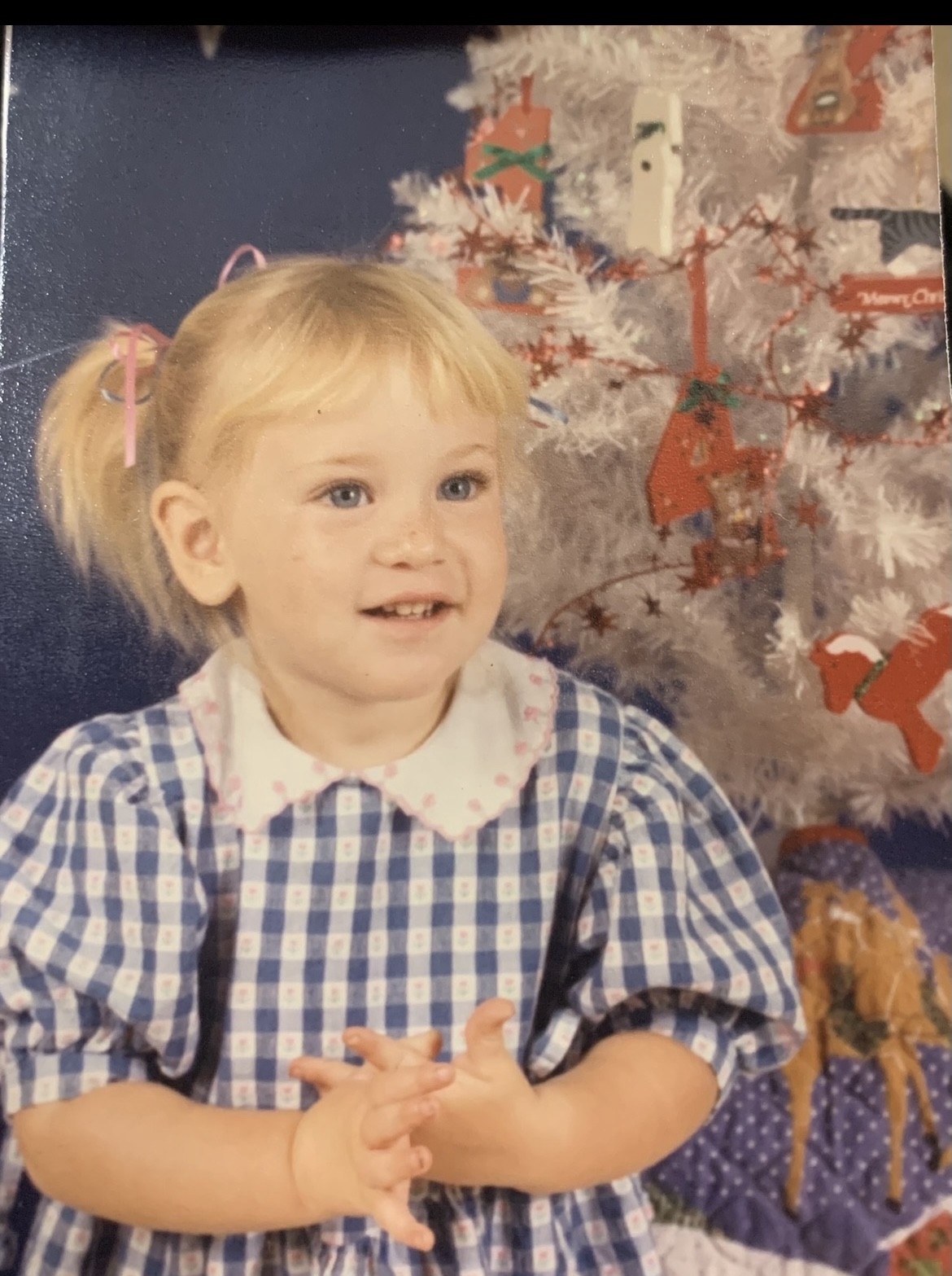 Ashlee as a toddler, blonde pigtails, blue gingham dress, white Christmas tree behind